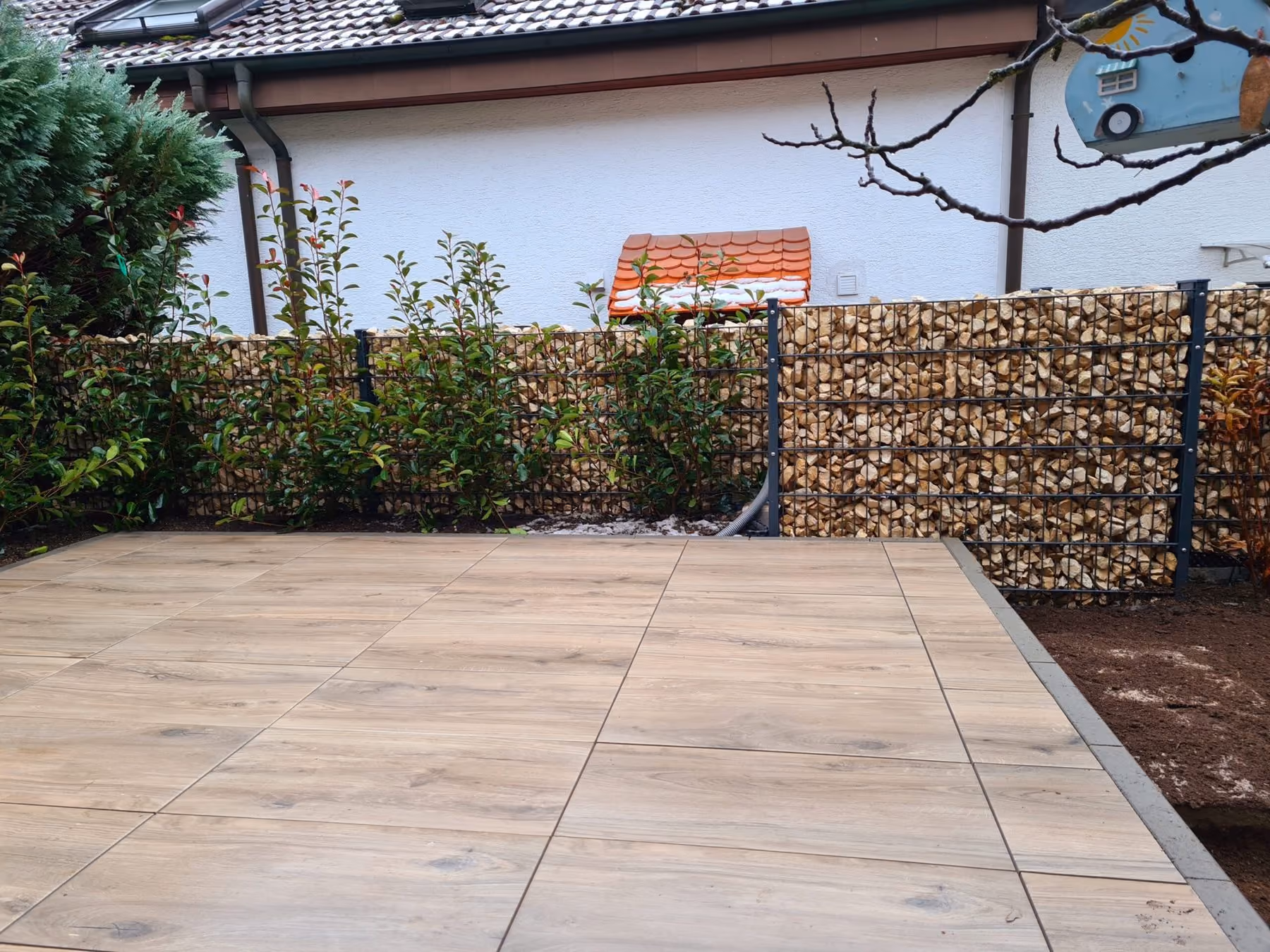 Terrasse aus hellen Großformatplatten, seitlich begrenzt von einer Gabionenwand mit Bepflanzung