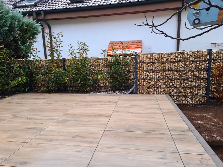 Terrasse aus hellen Großformatplatten, seitlich begrenzt von einer Gabionenwand mit Bepflanzung
