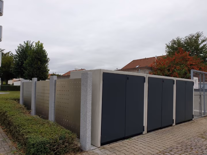 Müllplatzeinhausung mit anthrazitfarbenen Türen und hellen Betonpfosten an einer Hecke