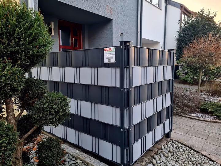 Müllplatzeinhausung mit anthrazitfarbenen Sichtschutzstreifen neben einem Hauseingang