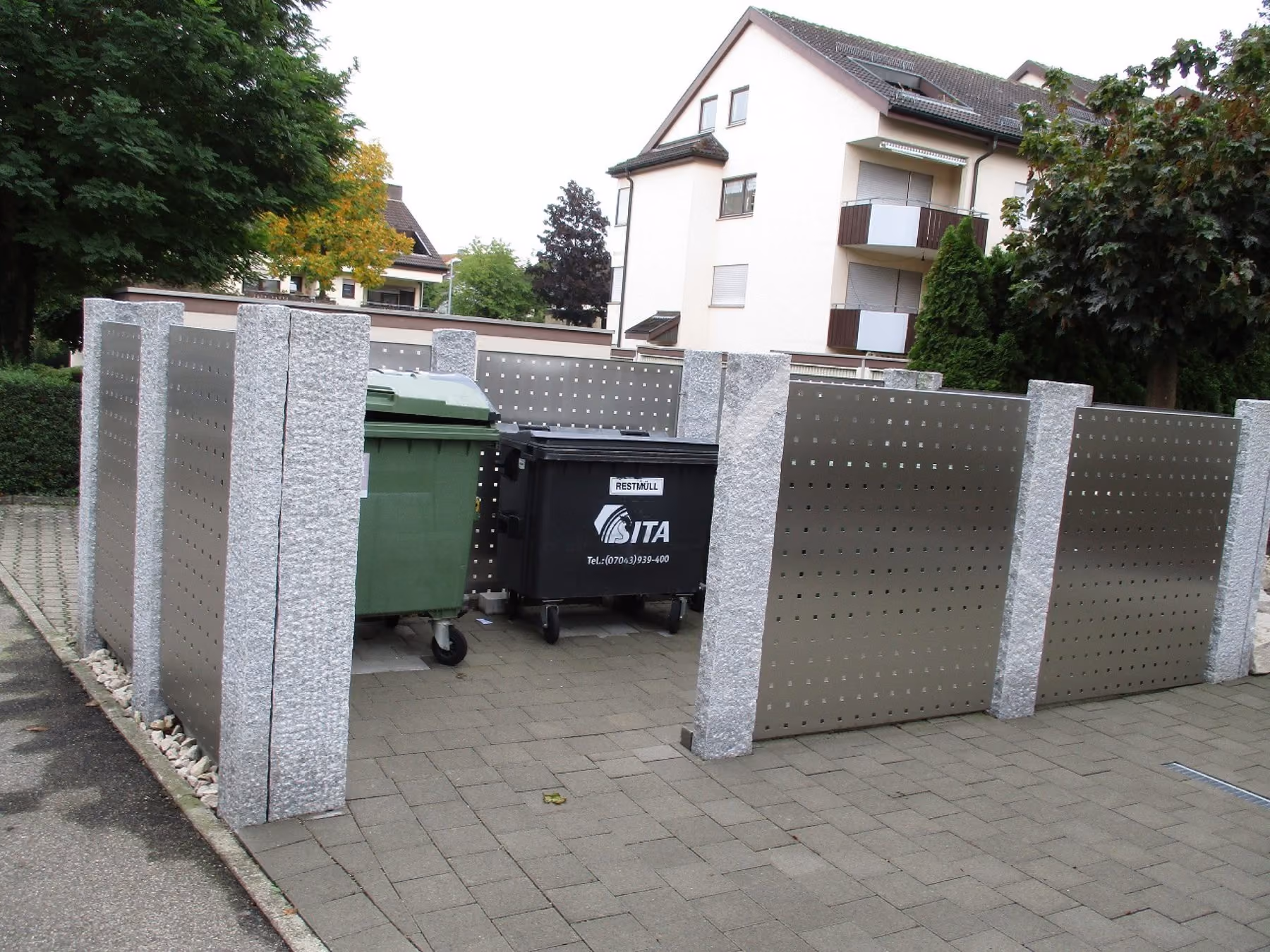 Müllplatz mit Edelstahl-Sichtschutzelementen zwischen Granitpfosten vor einem Wohnhaus