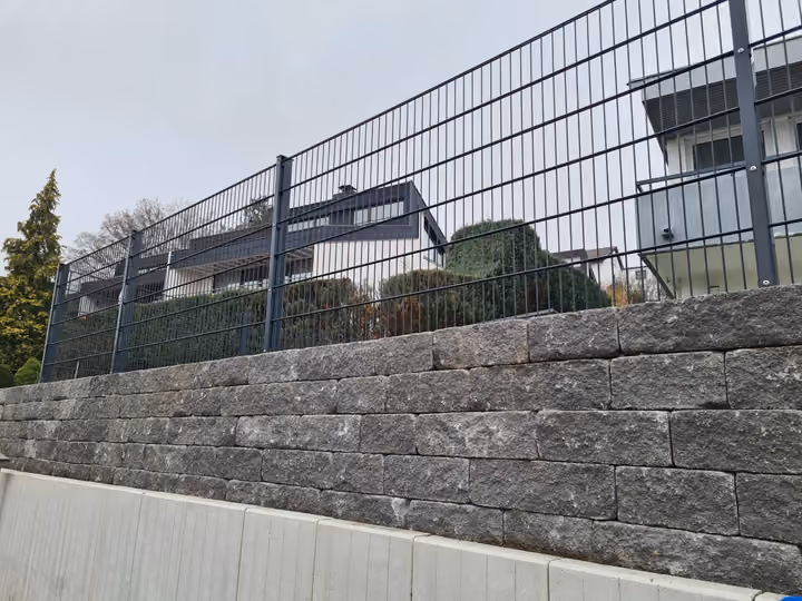 Trockenmauer aus geschichtetem Naturstein unterhalb eines Doppelstabmattenzauns