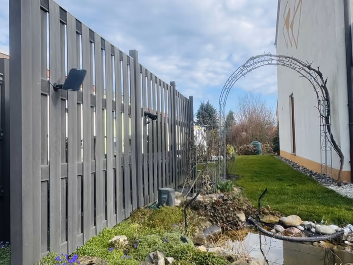 Hohe Sichtschutzwand entlang eines Gartenteichs mit blühender Uferbepflanzung