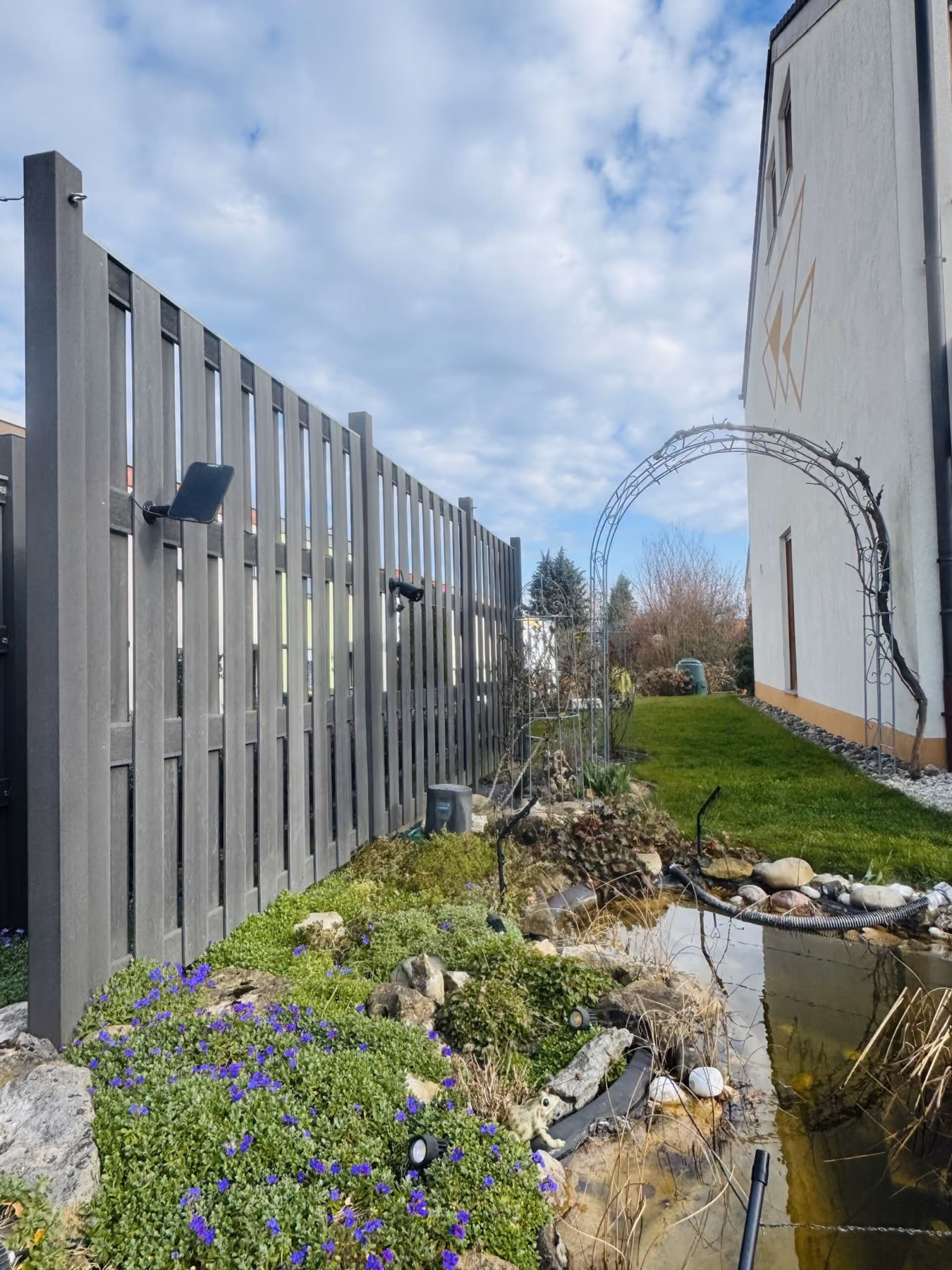 Hohe Sichtschutzwand entlang eines Gartenteichs mit blühender Uferbepflanzung